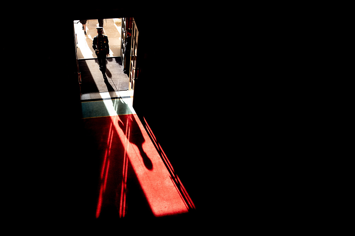 Image shows shadows and cross cast on the ground in front of silhouette of a RAF aviator as they enter into a dark building.